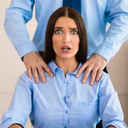 woman in blue shirt with dark hair has a shocked or worried look on her face when a man puts both his hands on her shoulders.