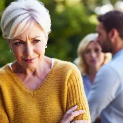 Older mother in yellow jumper with arms crossed with son and his partner in background.