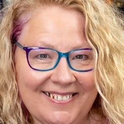 Jennifer, professional psychic reader with long blonde wavy hair and colourful blue and purple glasses, smiling warmly in a close-up portrait.