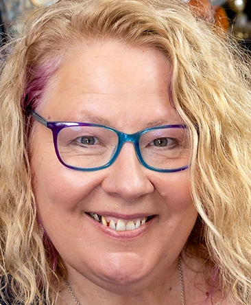 Jennifer, professional psychic reader with long blonde wavy hair and colourful blue and purple glasses, smiling warmly in a close-up portrait.